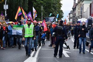 Zdjęcie kolorowe. Po lewej stronie grupa osób biorąca udział w marszu, po prawej stronie umundurowani policjanci oraz oznakowane policyjne radiowozy
