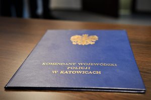 Zdjęcie kolorowe: granatowa teczka ze złotym napisem o treści Komendant Wojewódzki Policji w Katowicach