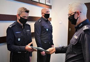 Zdjęcie kolorowe: dwóch umundurowanych policjantów stoi w szeregu, na przeciwko pierwszego z nich stoi umundurowany policjant i przekazuje mu teczkę