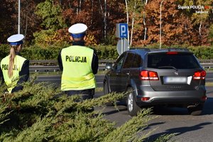 Policjanci - kobieta i mężczyzna podczas prowadzenia akcji Znicz w okolicy cmentarzy. Po prawej przejeżdża samochód