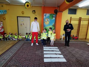 Olimpijczyk Dawid Tomala oraz policjant stoją przed ułożonym na wykładzinie w przedszkolu pasami imitującymi przejście dla pieszych i uczą dzieci jak prawidłowo przechodzić przez jezdnię.