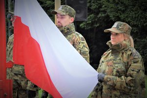 Kolorowe zdjęcie. Umundurowana kobieta trzyma flagę Polski, obok niej stoi umundurowany mężczyzna.