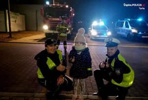 policjant i policjantka z dziewczynką pozują do zdjęcia. W tle strażak, wóz strażacki, radiowóz policyjny i straży miejskiej.