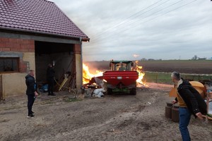 pożar drewnianych przedmiotów obok budynku, mężczyzna traktorem wjeżdża w płomienie, dwaj mężczyźni stoją i obserwują sytuację, trzeci mężczyzna wodą w węża ogrodowego gasi ogień