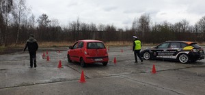Na fotografii widać funkcjonariusza policji oraz sędziego oceniających poprawność wykonania zadania przez kierującego samochodem osobowym marki Hyundai. Obok zaparkowany samochód rajdowy marki Subaru.