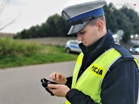 Na zdjęciu widoczny umundurowany policjant w odblaskowej kamizelce, trzymający urządzenie do sprawdzania danych osobowych.