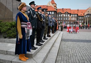 Reprezentanci różnych służb w mundurach oraz osoby w starodawnych strojach stoją w szeregu przed pomnikiem Wojciecha Korfantego.