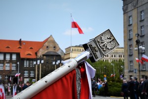 Zbliżenie na górną część sztandaru. Widoczny napis KWP Katowice i orzeł. W tle flaga Polski.