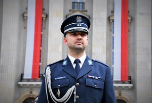 Policjant w mundurze galowym stojący na tle dwóch flag Polski.