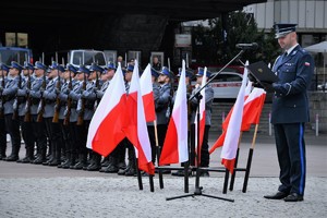 Umundurowany policjant odczytuje Apel Pamięci. Widoczna policyjna kompania honorowa i flagi Polski.