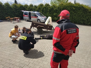 Na zdjęciu widać ratownika medycznego oraz strażaka, któzy egzaminują uczniów, któzy wykonują zadanie z pierwszej pomocy przedmedycznej.
