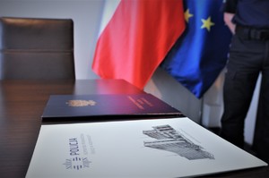 Teczki z rozkazami personalnymi leżą na biurku, w tle flaga Polski i Unii Europejskiej.