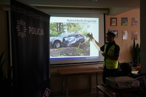 Na fotografii wiadać policjanta wydziału ruchu drogowego, który przezentuje zdjęcie zniszczonego pojazdu w wyniku zdarzenia drogowego.