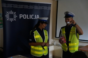 Na zdjęciu widac policnajtkę i policjanta wydziału ruchu drogowego. Policjant prezentuje element odblaskowy.