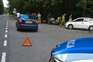 Zdjęcie z miejsca pozorowanego wypadku. Na fotografii widoczny pojazd, którego kierowca zatrzymał się celem udzielenia pomocy, trójkąt ostrzegawczy, a także policyjny radiowóz i strażacy.