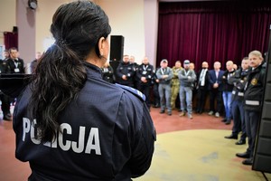 Na zdjęciu umundurowana policjantka. Widoczne jej plecy i napis policja, w tle przedstawiciele różnych służb oraz zaproszeni goście stojący w półokręgu.