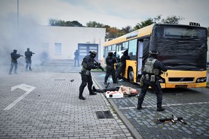 Zdjęcie. Widoczni umundurowani policjanci z bronią maszynową przed autobusem, oraz osoba leżąca na chodniku