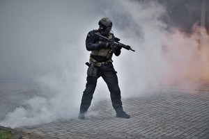Zdjęcie. Widoczny umundurowany policjant, granaty dymne, w tle wóz strazy pożarnej