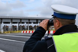 Na zdjęciu widzimy w tle bramki na autostradzie A4, na pierwszym planie jest widoczny umundurowany policjant w kamizelce odblaskowej patrzący przez lornetkę.