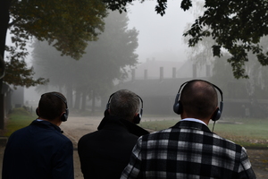 trzy osoby stojące tyłem. Na głowach osób słuchawki. W tle budynek za mgłą