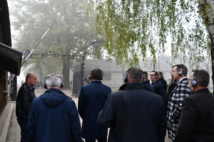 Osoby stojące w grupie i słuchające przewodnika. W tle brama obozu Auschwitz.