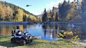 Zdjęcie. Widoczne jezioro, zaparkowany quad oraz śmigłowiec w locie