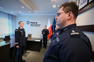 Zdjęcie przedstawia policjanta stojącego w szeregu, w tle znajduje się Komendant i policjant odczytujący rozkaz.