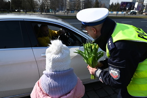 zdjęcie kolorowe przedstawiające policjanta z bukietem kwiatów i dziewczynkę przy białym samochodzie w którym widać mężczyznę za kierownicą