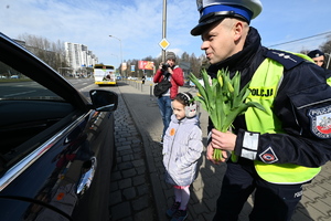 zdjęcie kolorowe przedstawiające policjanta z drogówki trzymającego bukiet kwiatów i dziewczynkę przy drzwiach samochodu, rozmawiają z kierowcą