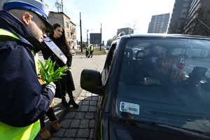 zdjęcie kolorowe przedstawiające policjanta z drogówki z kwiatami i Miss Polski przy samochodzie, w środku widać za kierownicą kobietę
