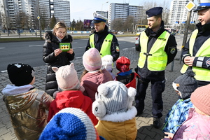 zdjęcie kolorowe przedstawiające policjantów i przedszkolaków na chodniku oraz kobietę czytającą kartkę z życzeniami z okazji 8 marca