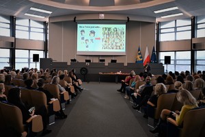 zdjęcie kolorowe w auli komendy, siedzące po obu stronach kobiety, przemawiający po prawej prelegent, na ekranie slajd z prezentacji