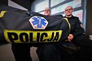 zdjęcie kolorowe w auli komendy, dwóch policjantów, jeden z torbą z emblematem ratownictwa medycznego i napisem policja