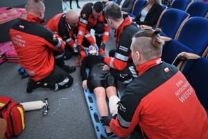 zdjęcie kolorowe w auli komendy, pokaz pierwszej pomocy, 5 ratowników przy desce ratowniczej na której leży kobieta