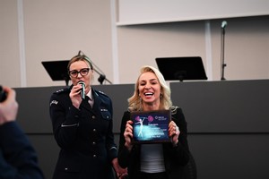 zdjęcie kolorowe w auli komendy, policjantka i kobieta z poprzedniej fotografii trzymająca okolicznościową tabliczkę