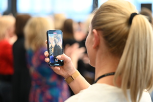 zdjęcie kolorowe w auli komendy, stojąca z telefonem kobieta, nagrywająca wydarzenie