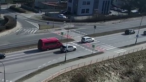 ujęcie z drona. Droga o dwóch pasach ruchu w jednym kierunku. Przed przejściem dla pieszych czerwony pojazd dostawczy, a z jego prawej strony srebrny mercedes.