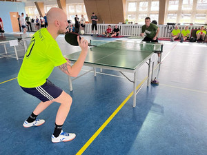 Na zdjęciu widać zawodników podczas rozgrywki tenisa stołowego. W tle widać innych zawodników.