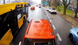 Zdjęcie z kamery samochodowej. Na zdjęciu widzimy czerwony samochód osobowy, którego kierowca przez okno wystawił rękę. Z lewej strony samochodu przejeżdża autobus.