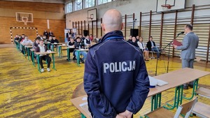 Zdjęcie wykonane w sali gimnastycznej. Na zdjęciu widzimy policjanta, a w tle uczniowie siedzący w ławkach.