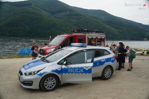 Na zdjęciu radiowóz policyjny i wóz strażacki nad jeziorem obok stoją dzieci.