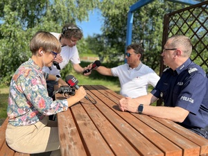 osoby siedzą na ławkach przy stoliku osoby z radia komendant wojewódzki oraz prezes radia jedna z osób podaje mikrofon prezesowi