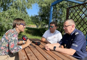 osoby siedzą na ławkach przy stoliku osoba z radia kobieta przeprowadza wywiad, na przeciw niej siedzą prezes radia oraz komendant wojewódzki