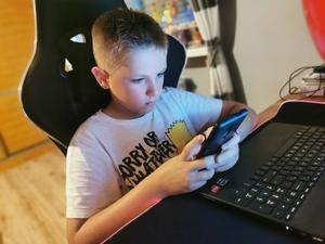 Na zdjęciu dziecko siedzi przed komputerem z telefonem w rękach.