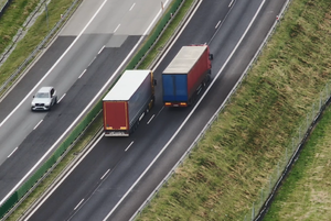 Zdjęcie przedstawia widok z drona. Na drodze widać dwa samochody ciężarowe jadące w tym samym kierunku oraz samochód osobowy jadący w kierunku przeciwnym.