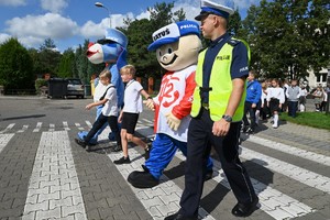 Zdjęcie. Umundurowany policjant, maskotka Policji i inna maskotka oraz dzieci przechodzące przez przejście dla pieszych
