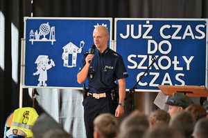 Zdjęcie. Widoczny umundurowany policjant z mikrofonem oraz dzieci na auli podczas uroczystości