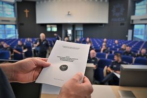 Na zdjęciu dłonie trzymają kartkę papieru, w tle policjanci przygotowujący się do testu z wiedzy.