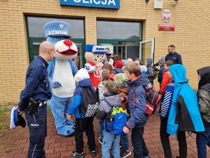 Policjant z maskotkami Sznupka i Ratusia witają dzieci przed wejściem do komisariatu