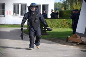 Zdjęcie. Policyjny pirotechnik w kostiumie sapera idzie droga, w tle umundurowani policjaci